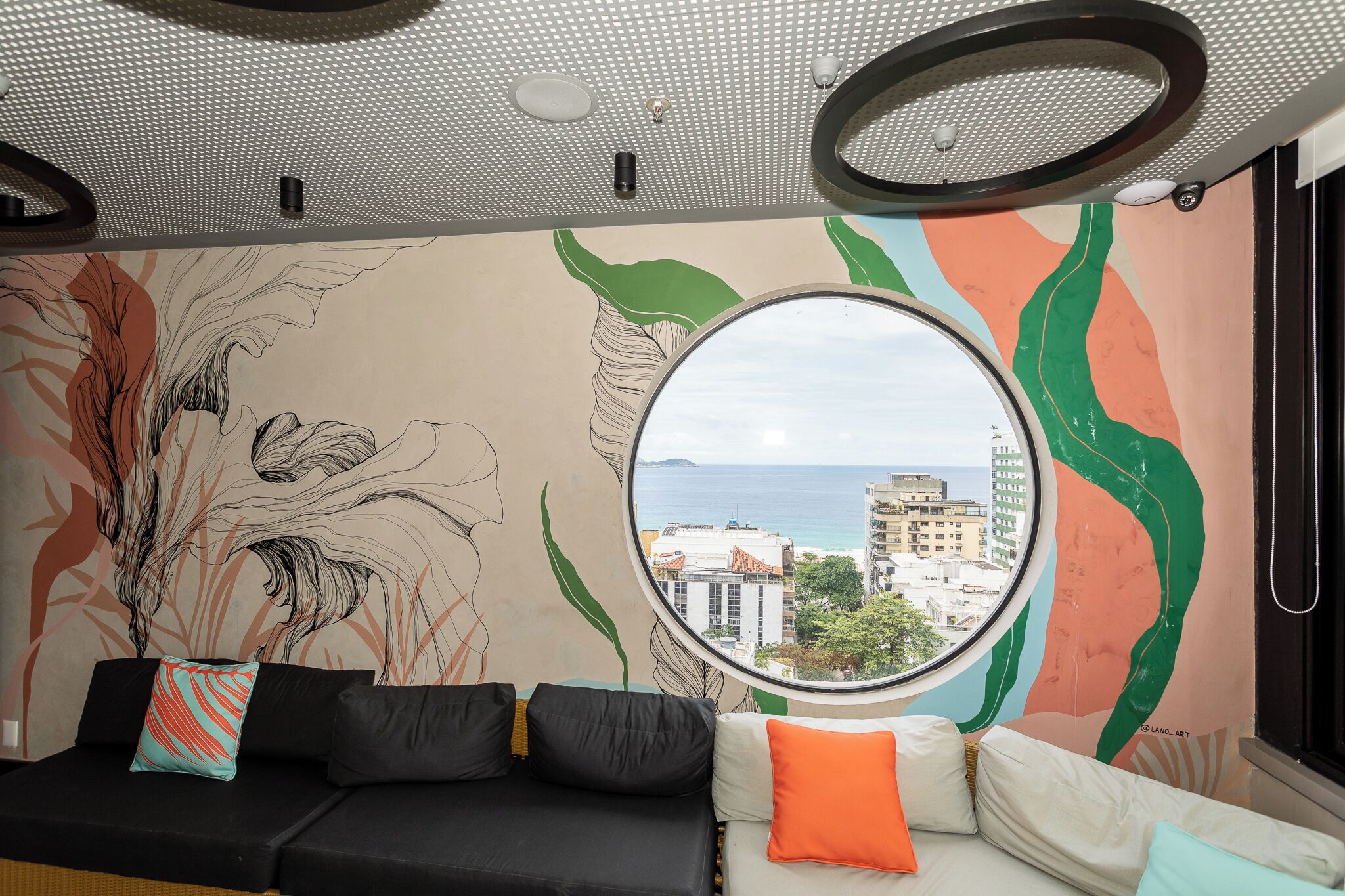 Habitació amb un sofà negre, un sofà blanc, un mirall rodó i un mural amb art abstracte i vista a la finestra.