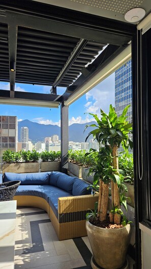 A balcony with a curved sofa, a large potted plant, and a view of a cityscape with mountains in the background. -  Hotel Astoria Ipanema (Rio de Janeiro)