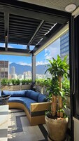 Varanda com sofá curvo, uma planta grande em vaso e vista para uma paisagem urbana com montanhas ao fundo.