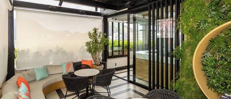 Un área de estar al aire libre moderna con sofá, sillas y una mesa, rodeada de vegetación y vistas a la ciudad.