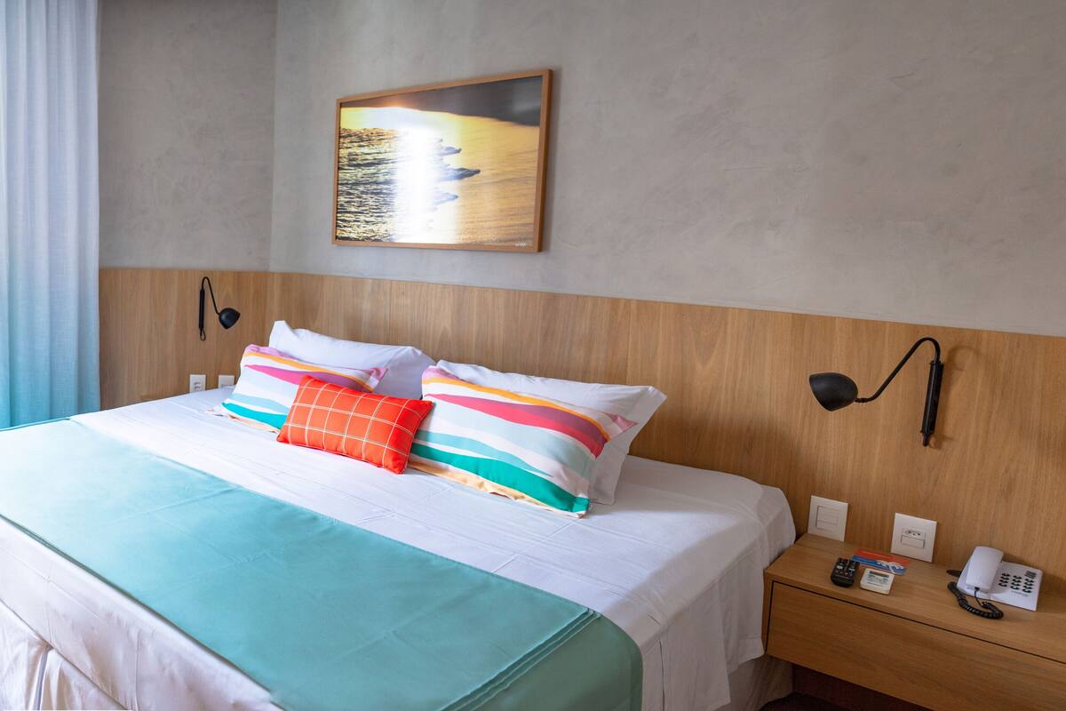Habitación de hotel con una cama grande, una mesita de noche con teléfono y lámpara, un cuadro en la pared y una ventana con cortinas.