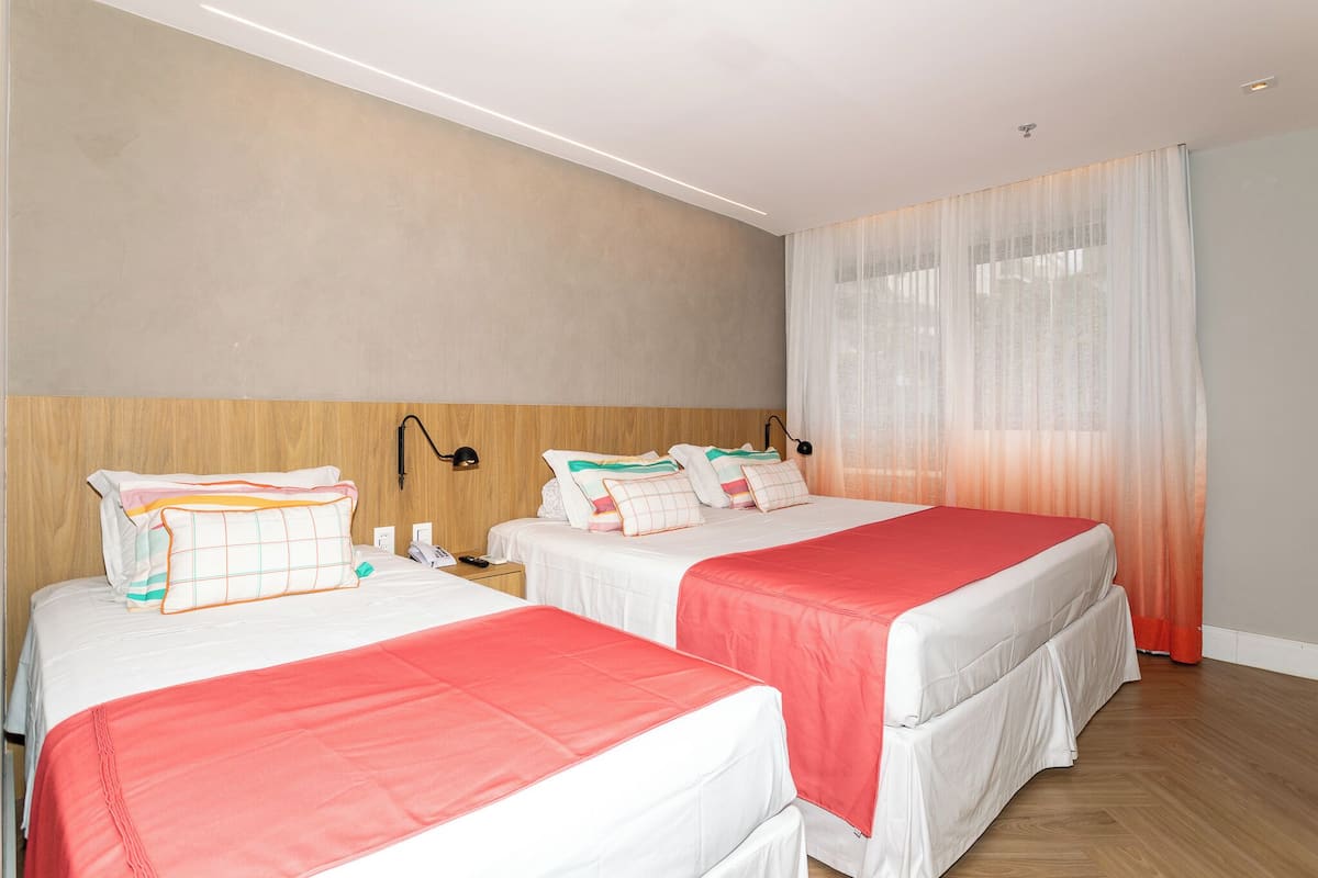 Habitación de hotel con dos camas, cabecera de madera, mesita de noche y ventana con cortinas traslúcidas.