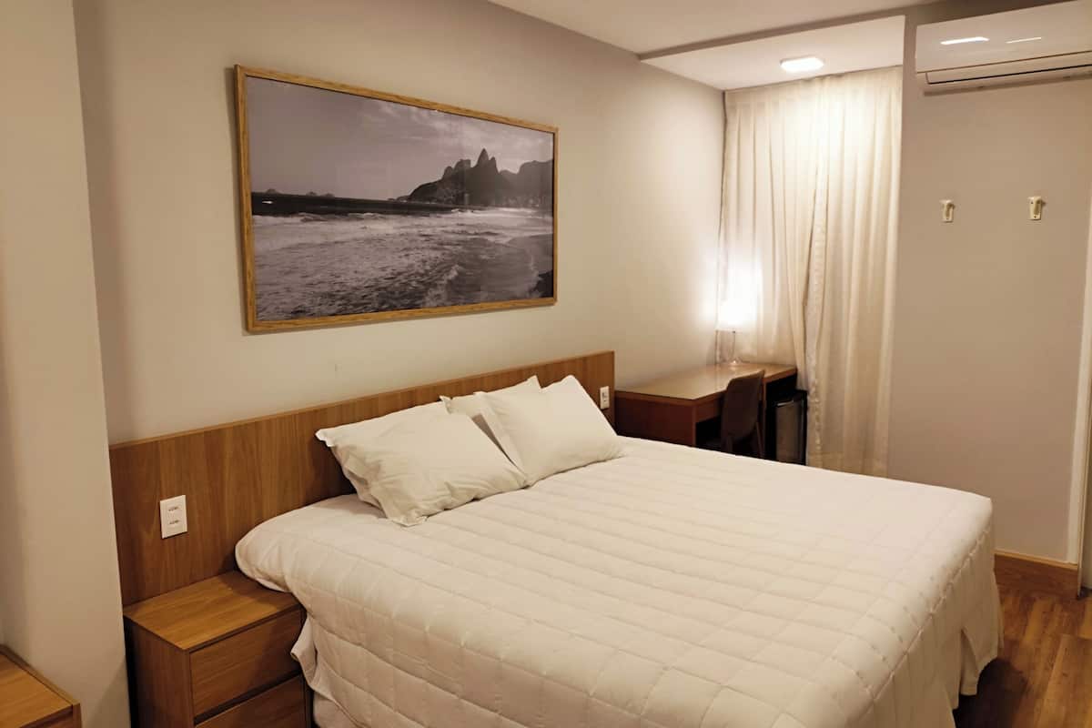 Habitación de hotel con cama, escritorio, silla, mesita de noche, un cuadro en la pared y aire acondicionado.