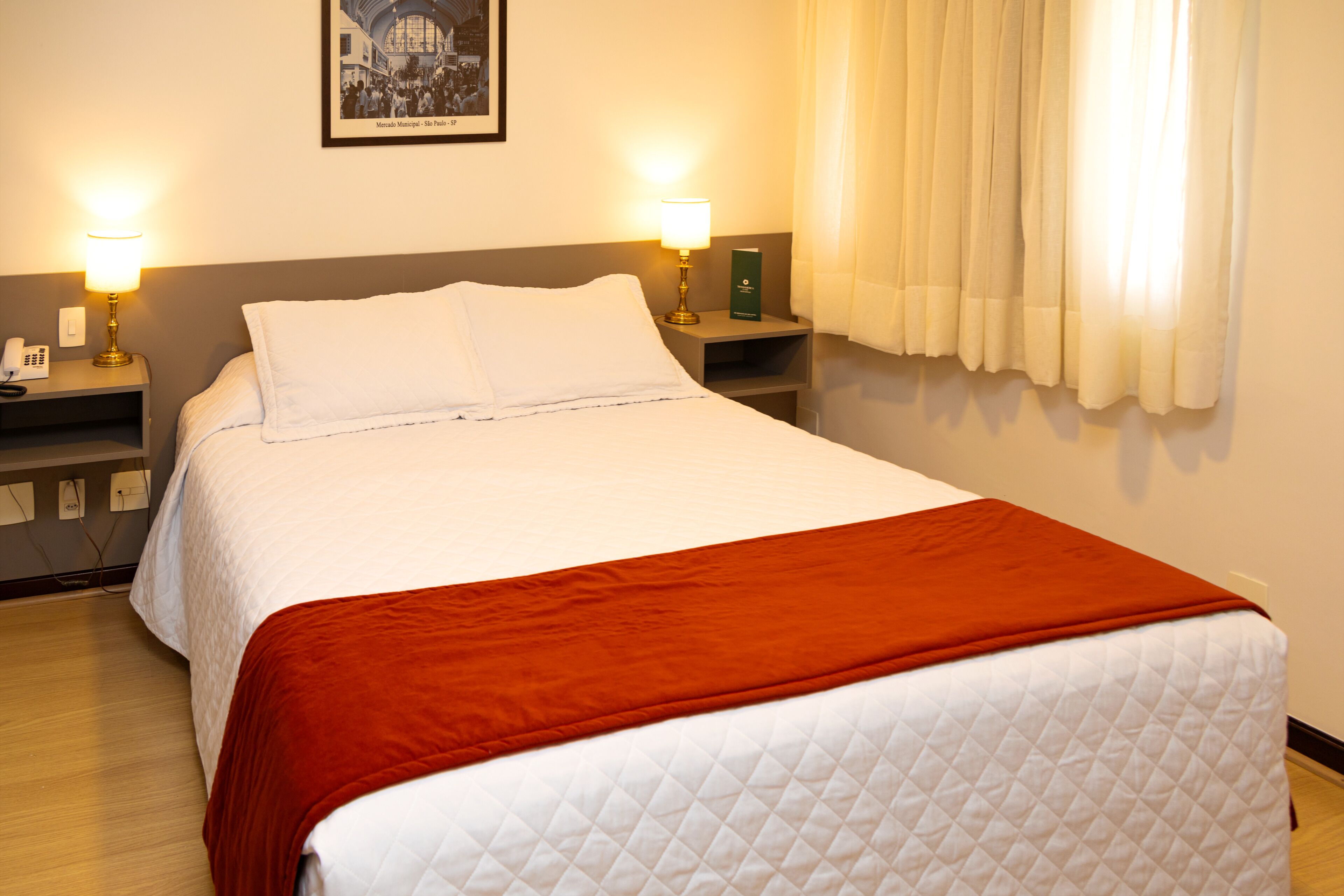 A neatly made bed with a white quilt and a red blanket, two bedside lamps, a framed picture on the wall, and a window with curtains.