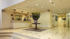 A hotel lobby with a reception desk, a vase with flowers, a staircase, and a polished floor.