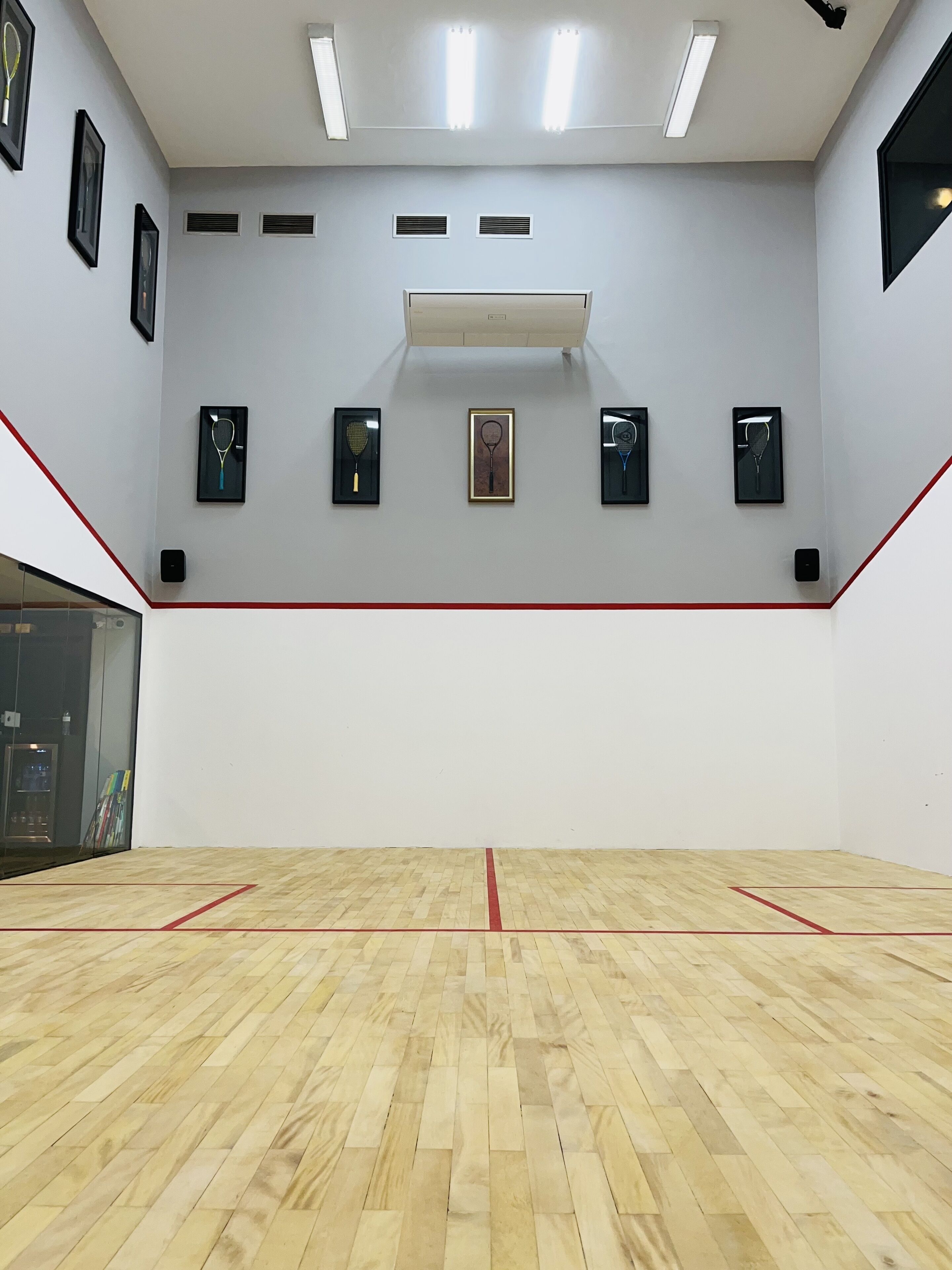 A squash court with a wooden floor, red boundary lines, and framed tennis rackets on the walls.