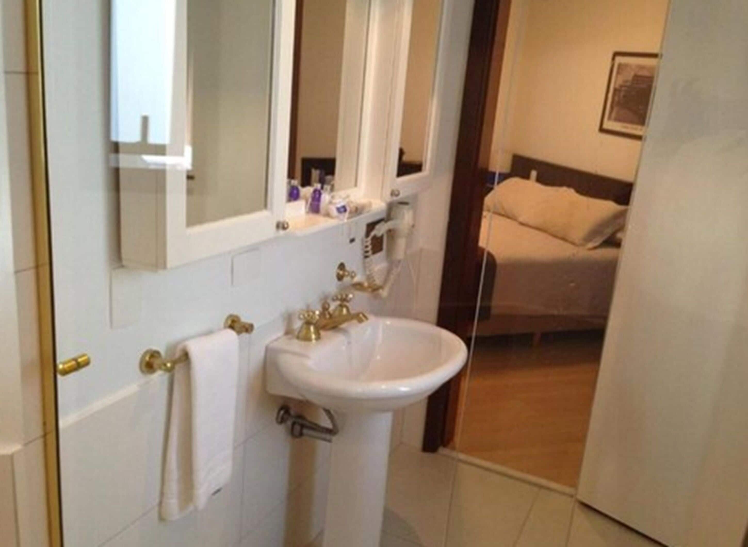 Un baño con lavamanos blanco, grifería dorada y un espejo encima. Hay una toalla colgada en la pared y una cama se ve al fondo.