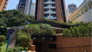 A tall, modern building with a central entrance, surrounded by greenery and other high-rise buildings.