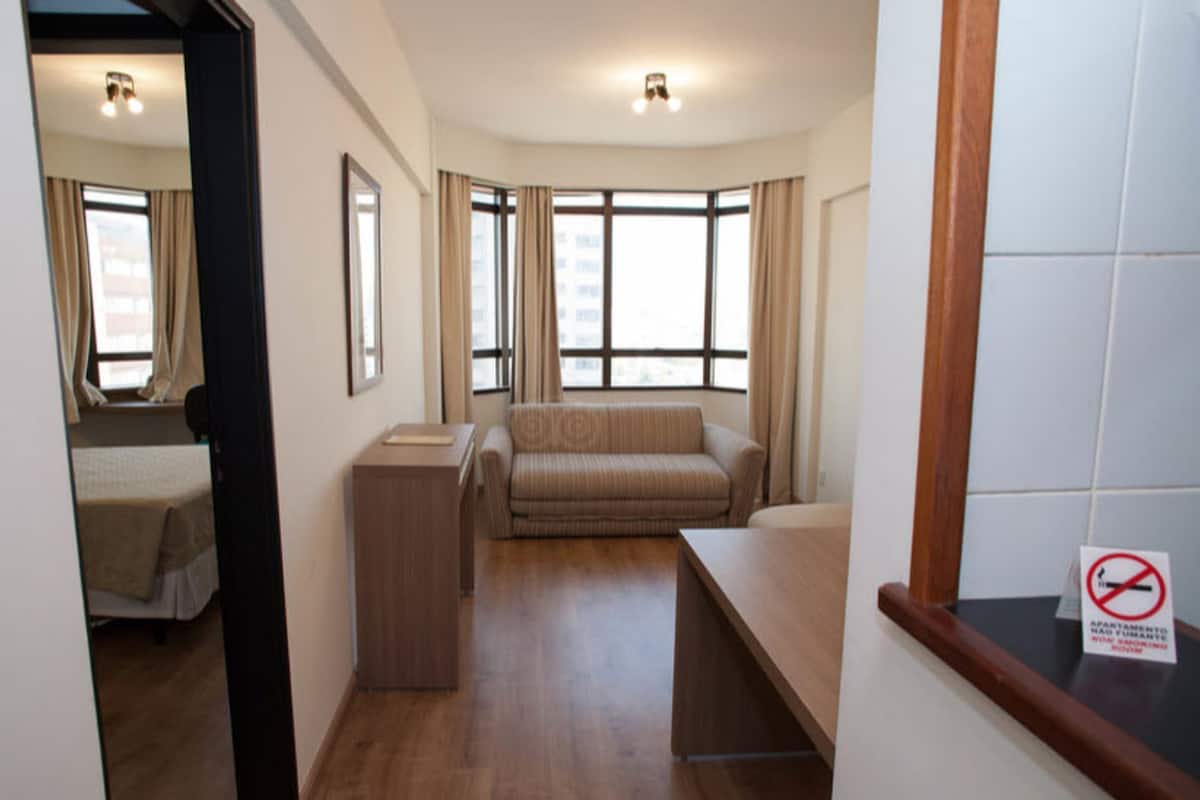 Une chambre d’hôtel avec un lit, un canapé, un bureau et une fenêtre avec des rideaux. Un panneau indique qu’il est interdit de fumer.