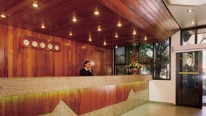 A reception area with a wooden reception desk, a clock displaying time, and a window with a view of greenery. - L'Hirondelle Flat Service (Campinas)
