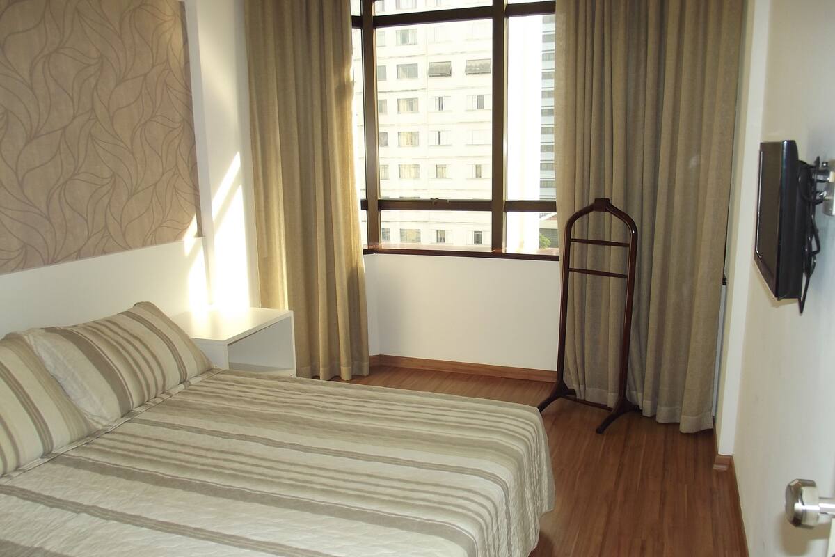 Une chambre d’hôtel avec un lit, une télévision, une fenêtre avec des rideaux et une vue sur les bâtiments environnants.