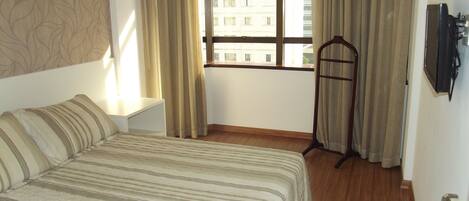 Habitación de hotel con cama, televisión, ventana con cortinas y vistas a edificios.
