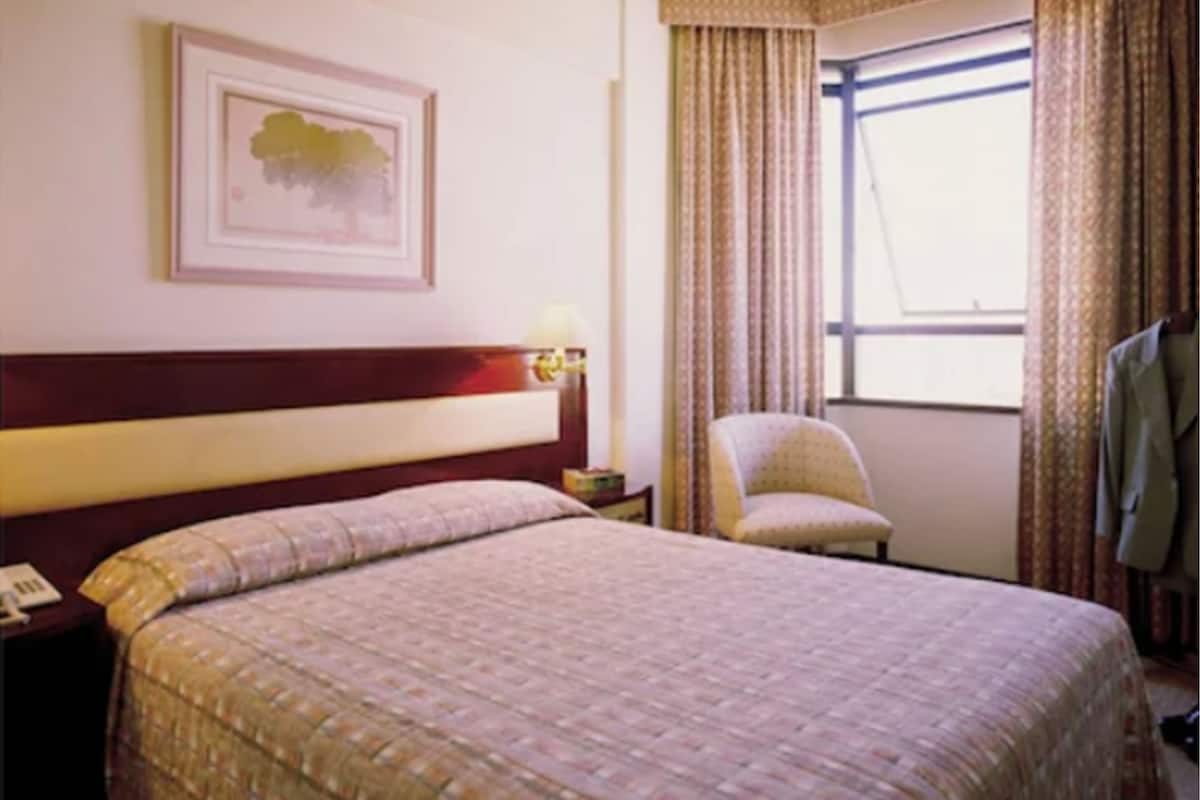 Une chambre d’hôtel avec un grand lit, une chaise, une table de chevet, un tableau encadré au mur et une fenêtre avec des rideaux.