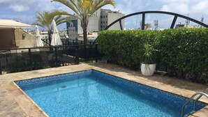 Una piscina rectangular con mosaicos azules, rodeada por un solárium de piedra y una barandilla de metal.