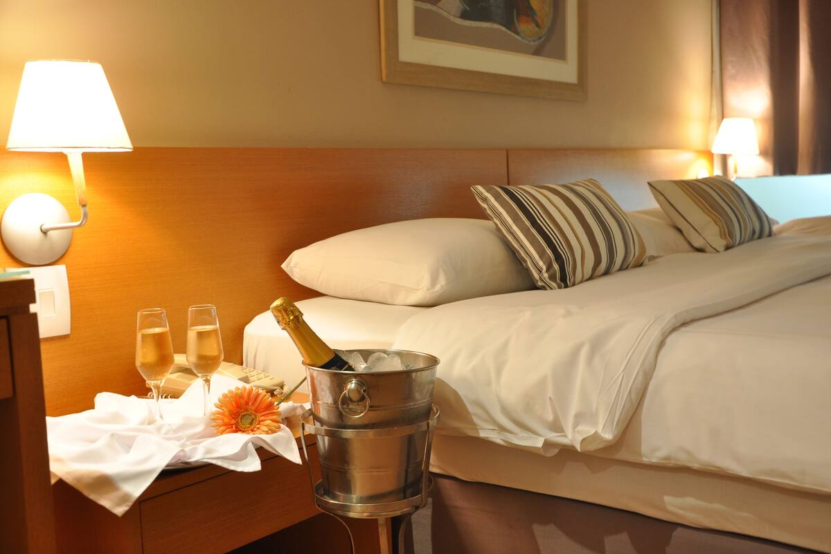 A hotel room with a bed, pillows, a nightstand with a lamp, a bucket with champagne, and glasses.