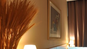 Una habitación de hotel con cama, lámpara de noche, un florero con plantas secas y un cuadro en la pared.