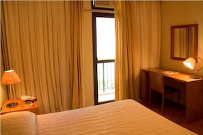 A hotel room with a bed, a desk, a chair, a lamp, and a window with curtains. - Promenade The Claridge (Rio De Janeiro)