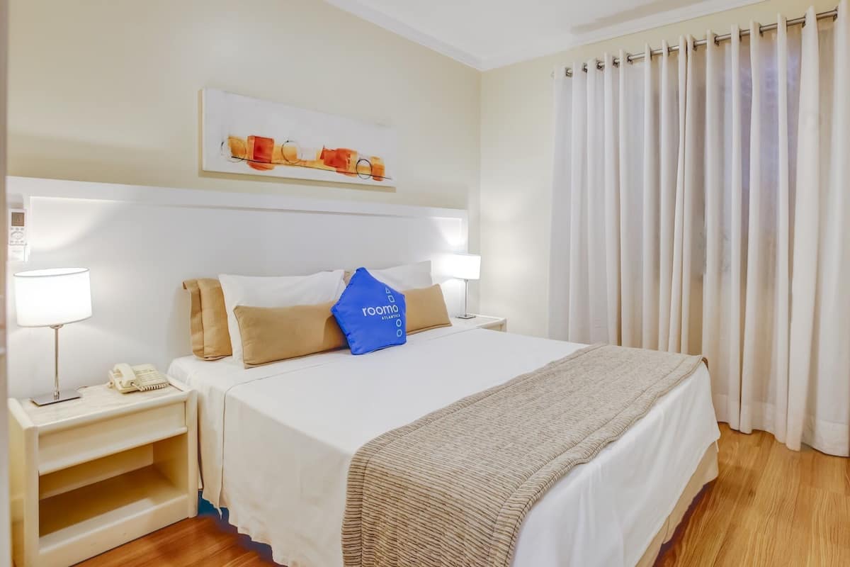A hotel room with a bed, two bedside tables, a lamp, a phone, a painting on the wall, and a curtain.