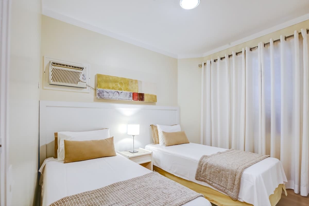 A hotel room with two beds, a bedside table with a lamp, a ceiling fan, and a window with curtains.