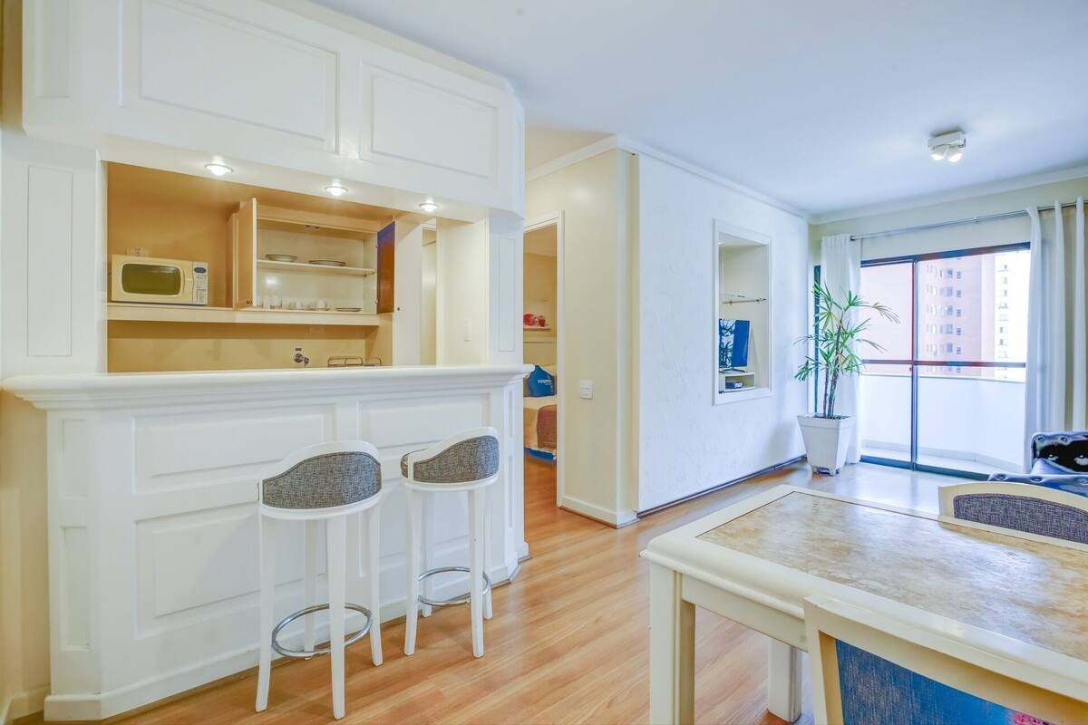 A modern kitchen with white cabinetry, a microwave, and two bar stools. There is a dining table with a blue accent and a potted plant by the window.