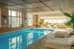 An indoor swimming pool with clear glass walls, a brick wall, and a ceiling with a grid pattern. There are lounge chairs and potted plants around the pool area. - InterContinental Santiago by IHG (Santiago)