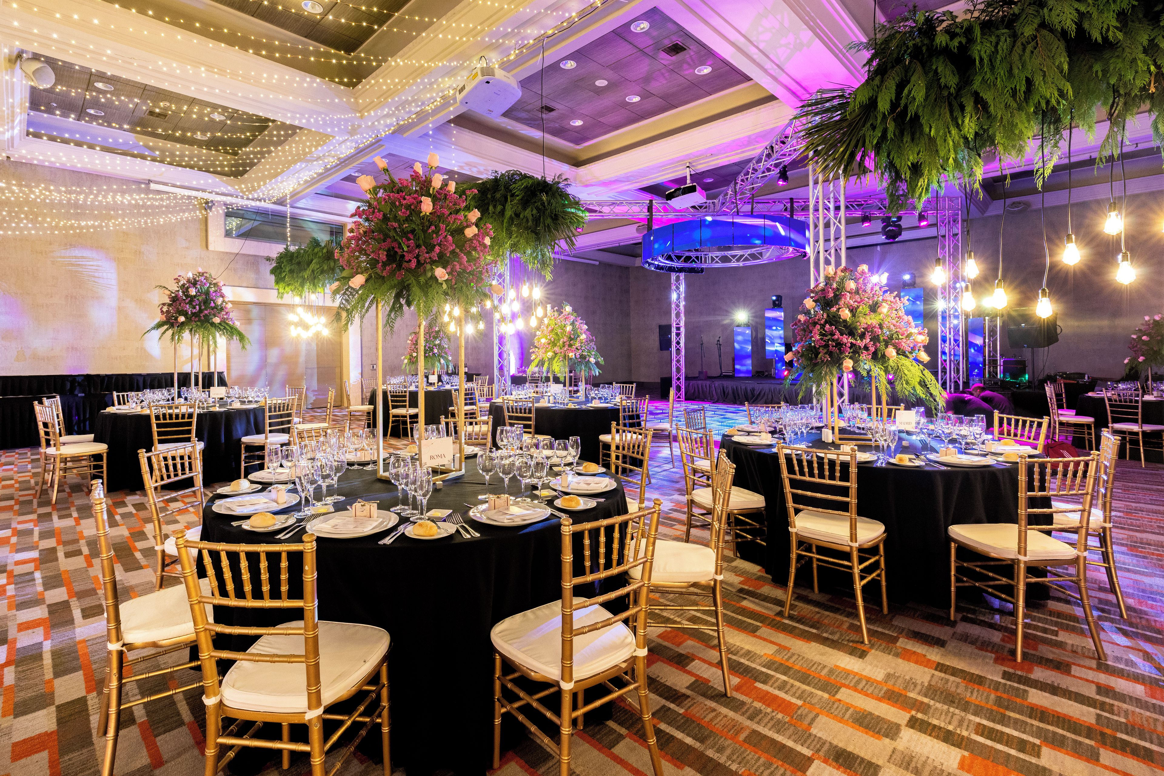 Un salón de banquetes con mesas redondas dispuestas para cenar, sillas doradas y centros de mesa con flores.
