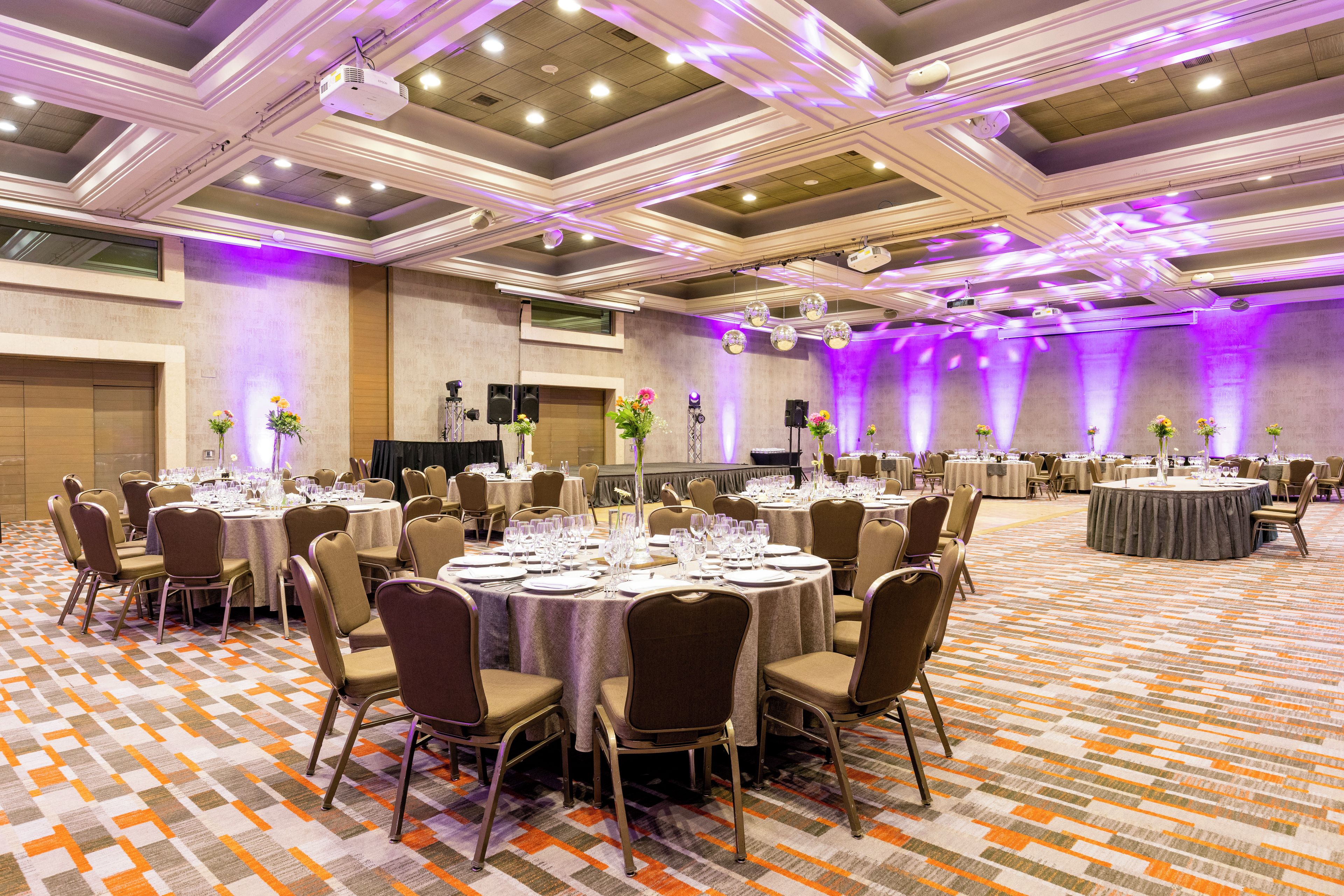 Un salón de banquetes con mesas redondas, sillas y un escenario con altavoces.