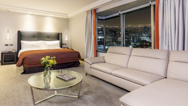 Ein Hotelzimmer mit Bett, einer Couch, einem Glastisch und Blick auf eine nächtliche Stadtlandschaft.