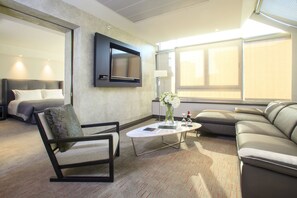 A modern hotel room with a large bed, a sofa, a chair, a coffee table, and a TV mounted on the wall.