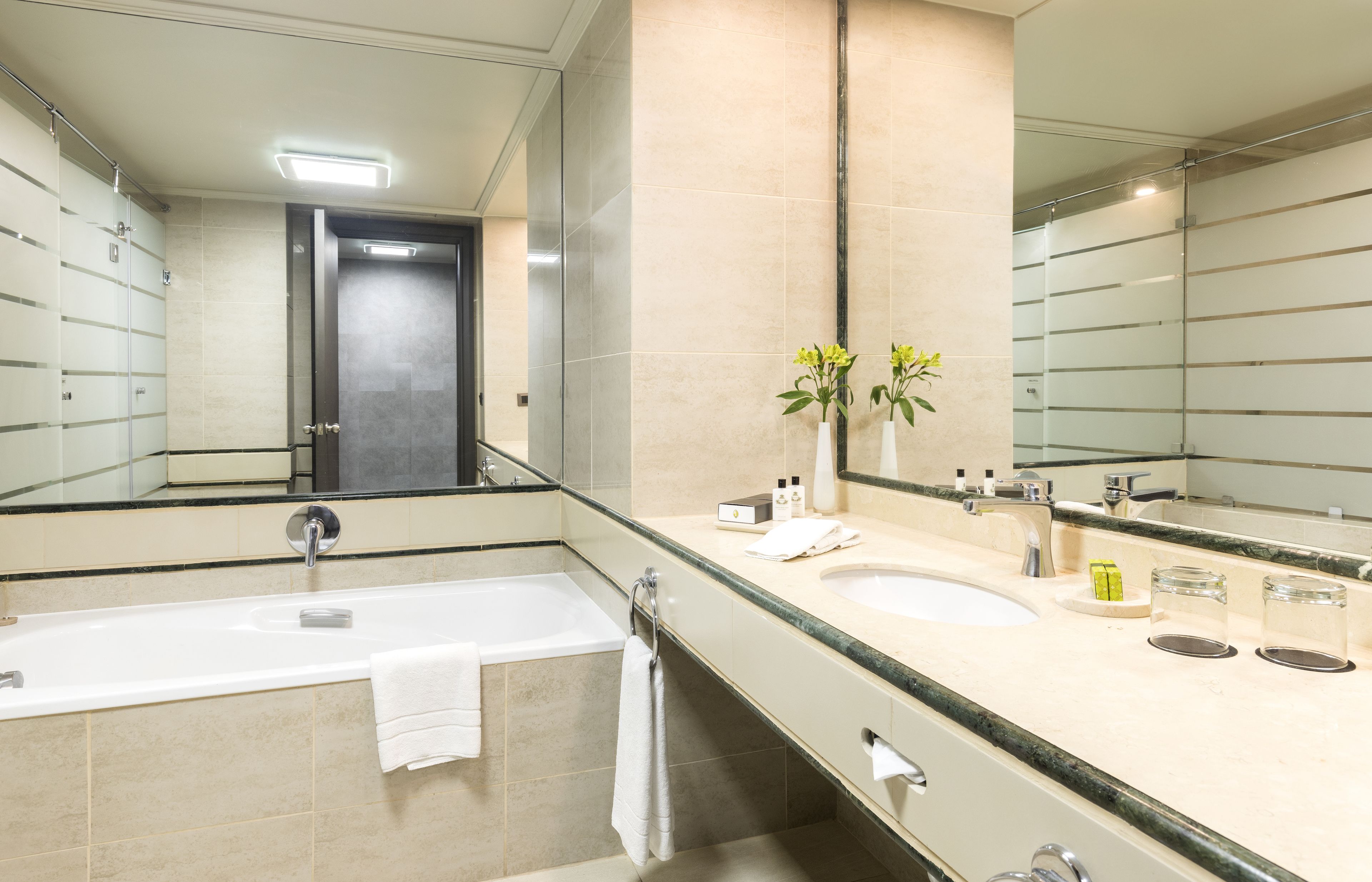A modern bathroom with a bathtub, two sinks, a large mirror, and a door leading to another room.