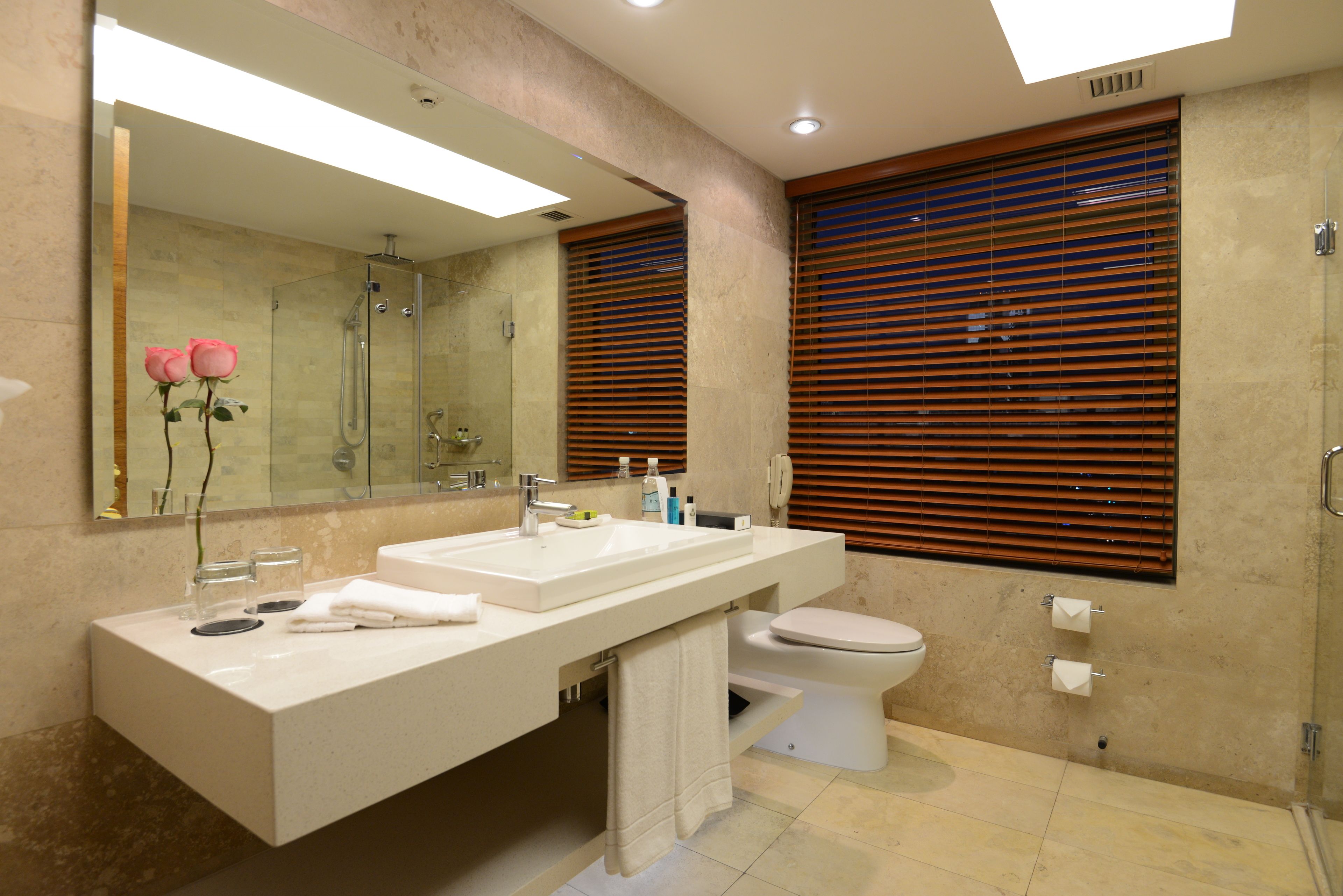 Ein modernes Badezimmer mit großem Spiegel, zwei Waschbecken, Toilette und Holzjalousien.