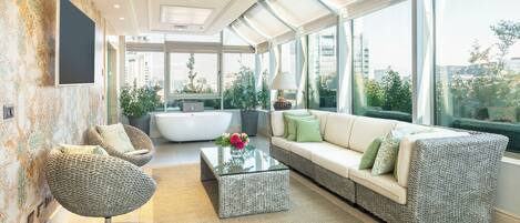 Un salon moderne comprenant un canapé, un fauteuil et une table basse, de grandes fenêtres offrant une vue sur la ville et un mur décoré d’un motif.