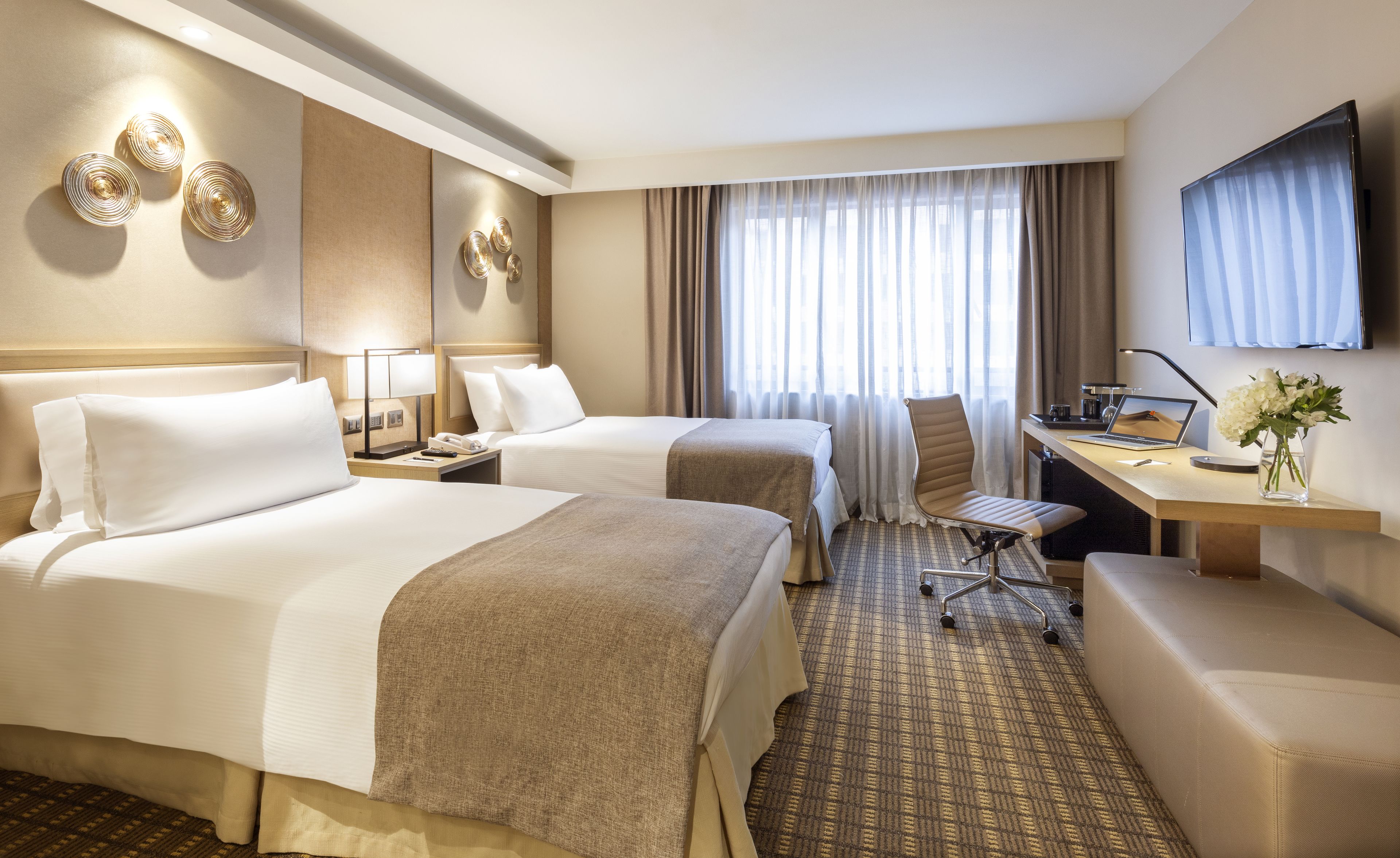 A hotel room with two beds, a desk, a chair, a TV, and a vase with flowers.