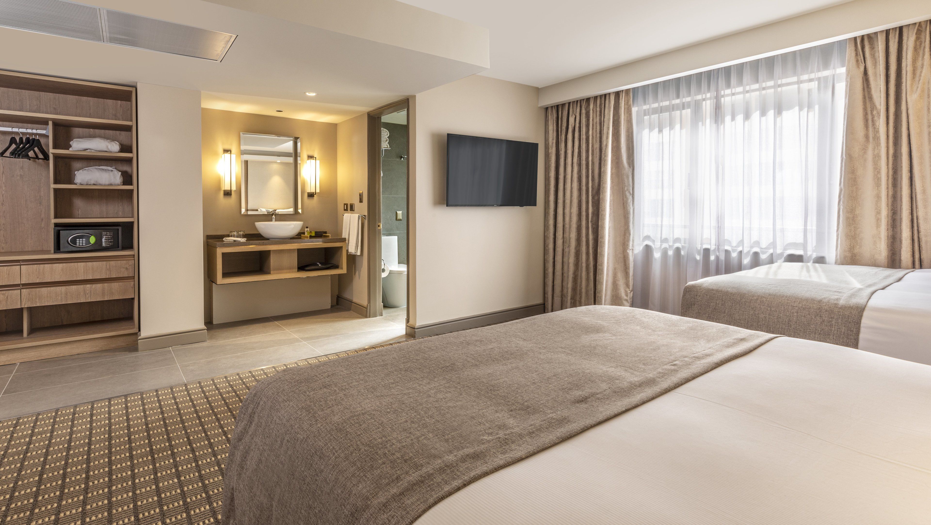 Habitación de hotel con cama grande, mesita de noche, televisor integrado, baño con lavamanos y espejo, y armario con ropa colgada.