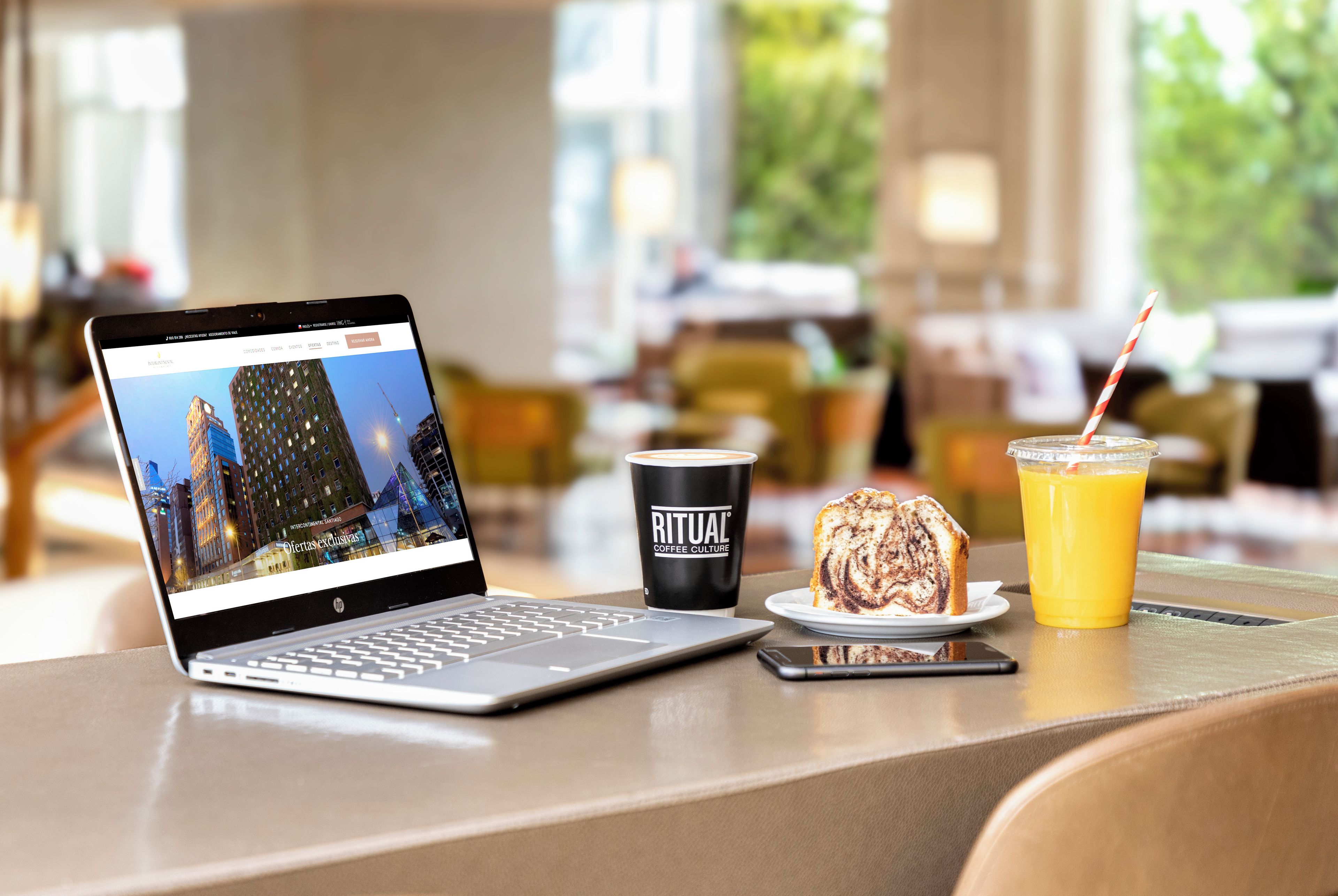 Una laptop mostrando un sitio web de hotel, una taza de café, una porción de pastel y un vaso de jugo de naranja sobre una mesa.