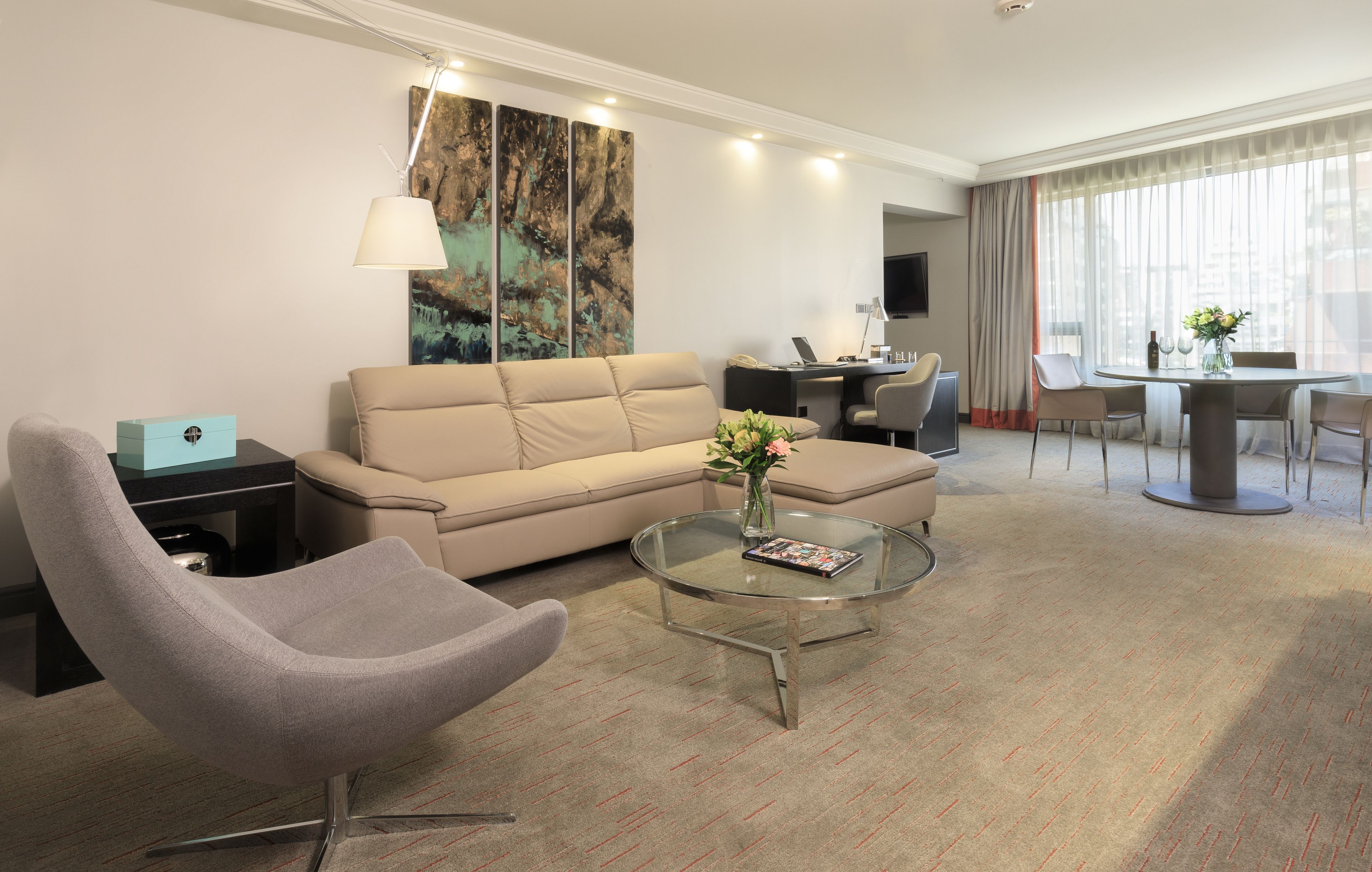 A modern hotel room with a sofa, armchair, glass coffee table, and a dining area with a table and chairs.