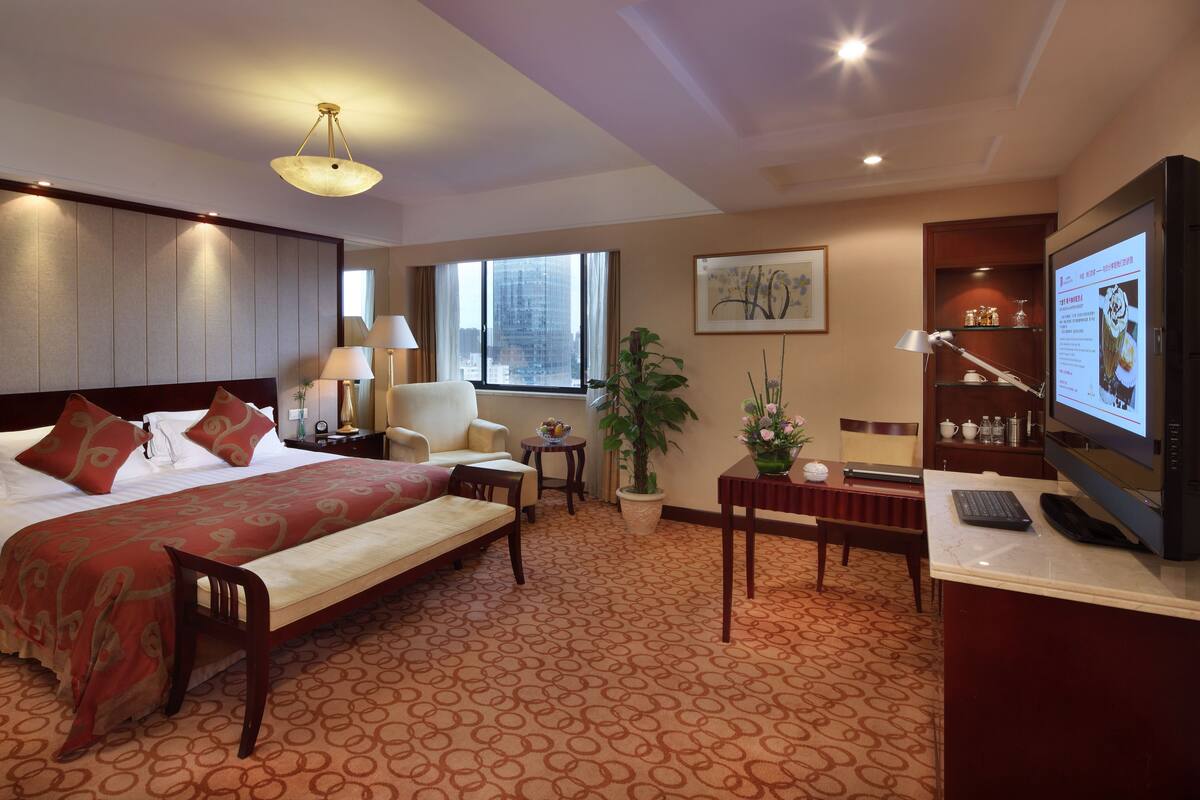 A hotel room with a bed, a desk with a computer, a chair, a small table, a lamp, a plant, and a television.