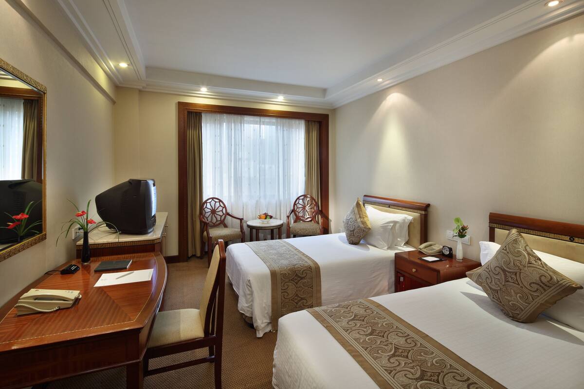A hotel room with two beds, a desk, a television, a chair, a small table, and a window with curtains.