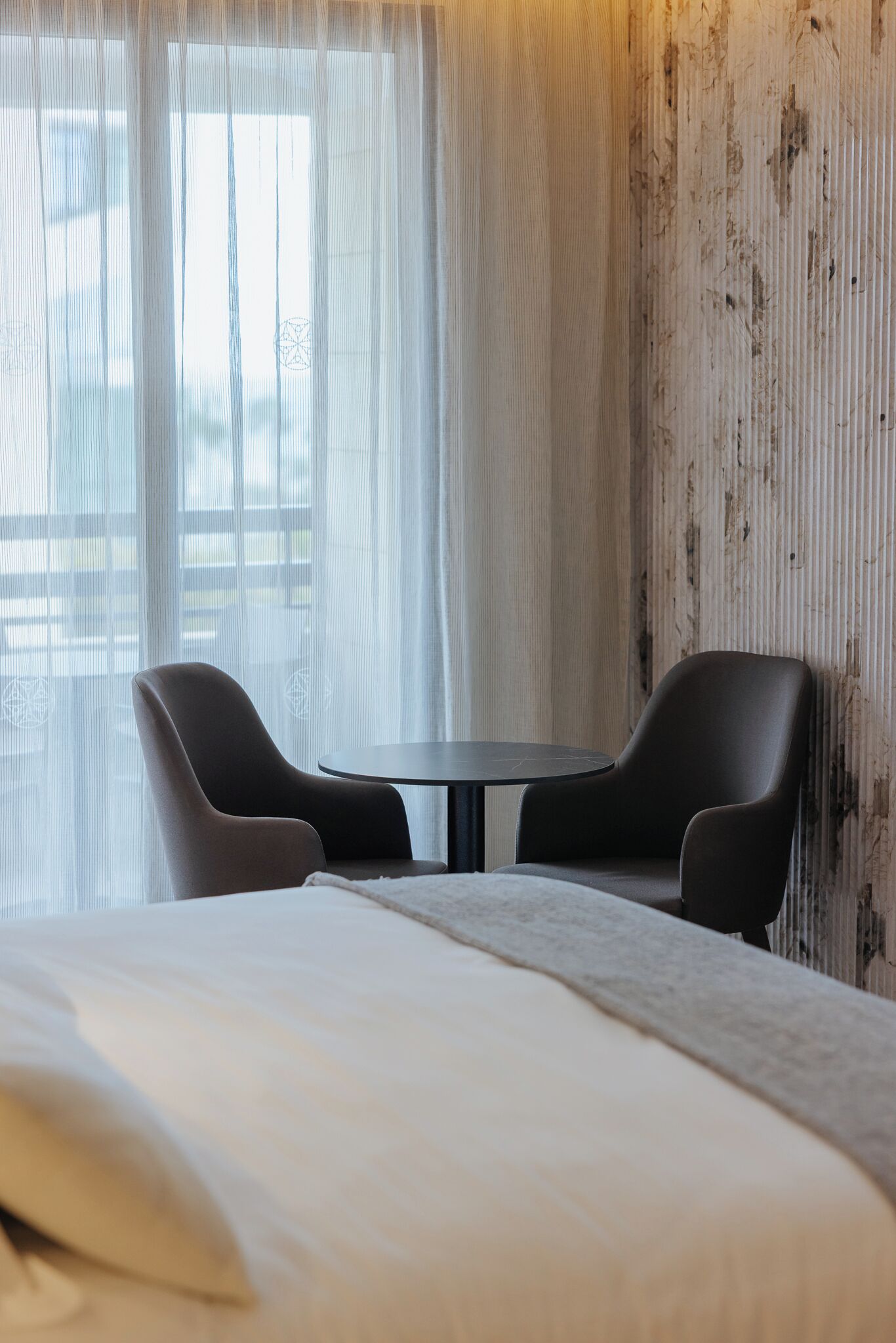 Une chambre d’hôtel avec un lit, deux chaises, une petite table et une fenêtre avec des rideaux légers.