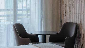 Quarto de hotel com cama, duas cadeiras, uma mesinha e janela com cortinas leves.