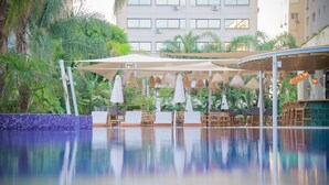Zona de piscina con sillas de descanso y sombrillas, rodeada de vegetación y un edificio al fondo.