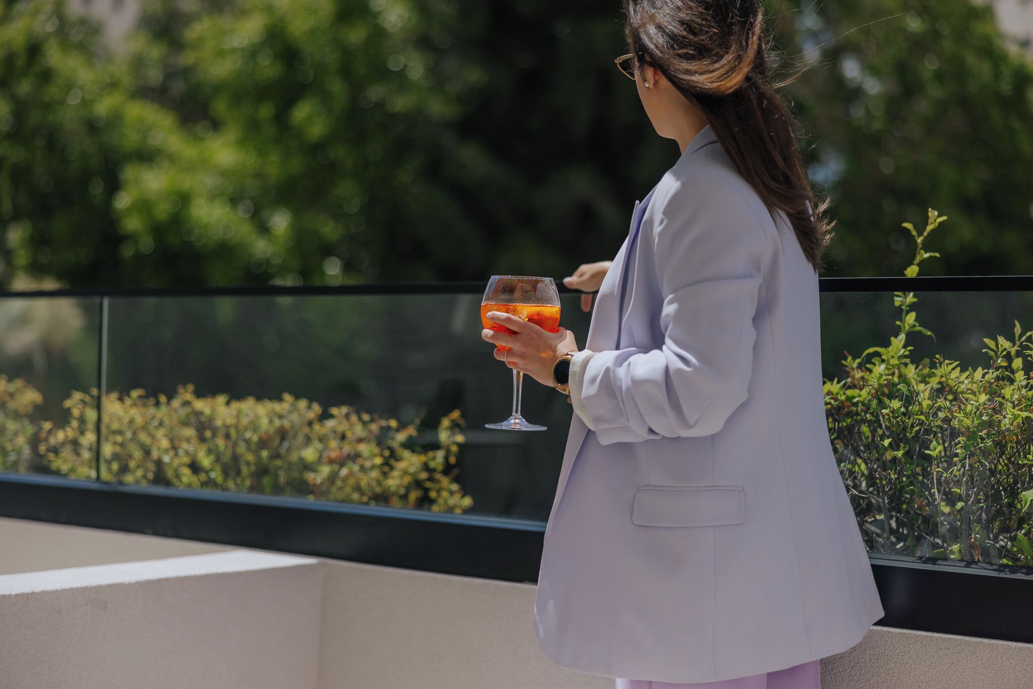 Una persona con un blazer de color claro sosteniendo un vaso con bebida, de pie en un balcón con vistas a zonas verdes y barandilla de cristal.