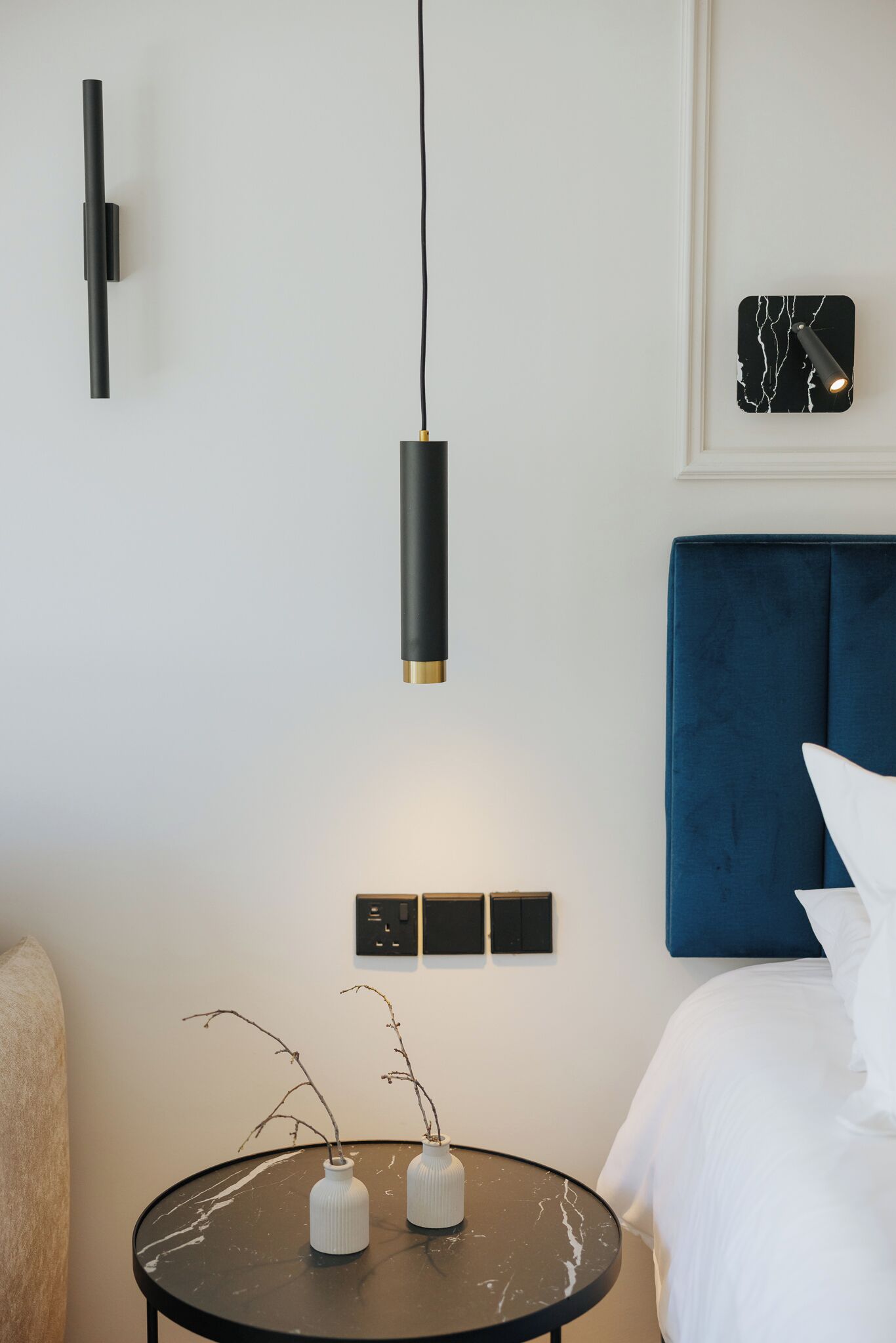 Quarto de hotel moderno com uma cama, uma mesa redonda com vasos, uma luminária na parede e um painel de cabeceira azul.