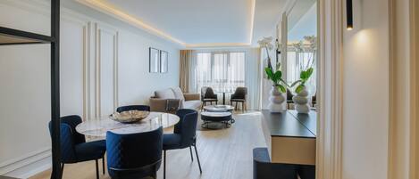 A modern living room with a sofa, two armchairs, a coffee table, and a dining table with chairs. There are potted plants on the windowsill and a large mirror on the wall.