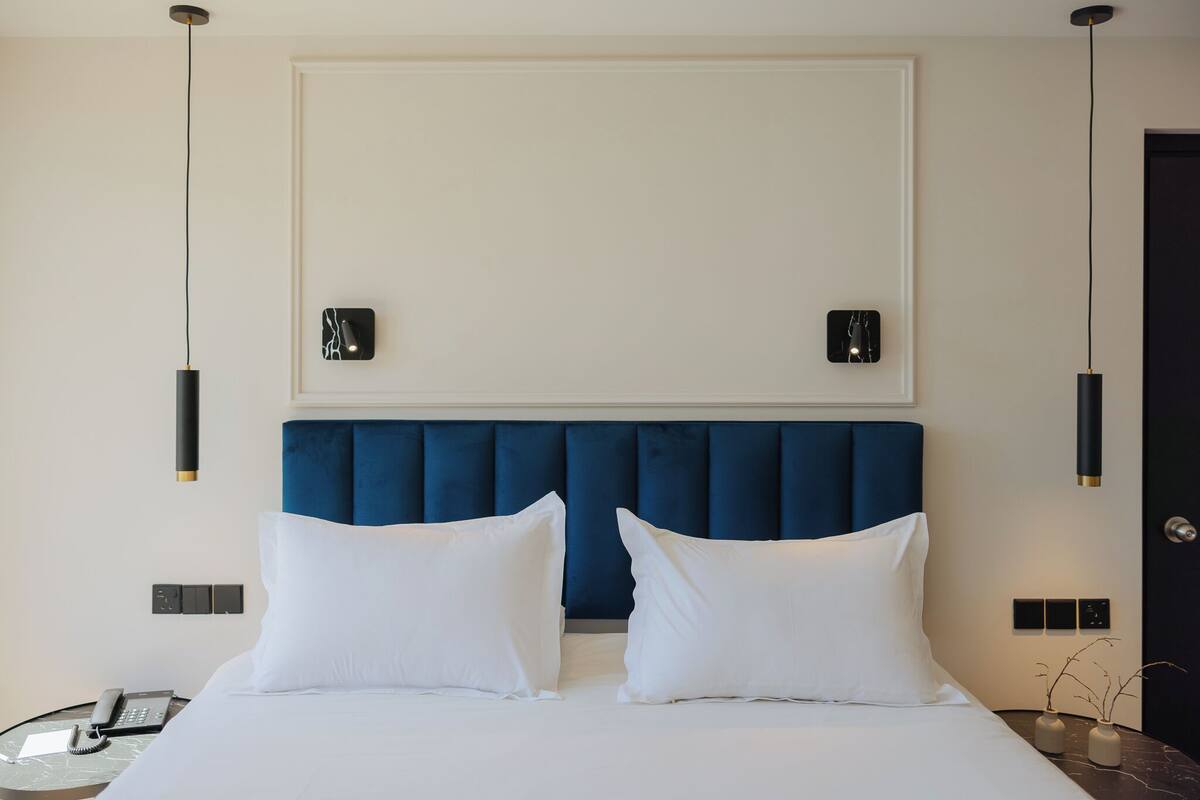 Ein Hotelzimmer mit einem großen Bett, einem blauen Stoffpolster-Kopfstück, zwei Nachttischen mit Lampen und einem an der Wand befestigten Telefon.