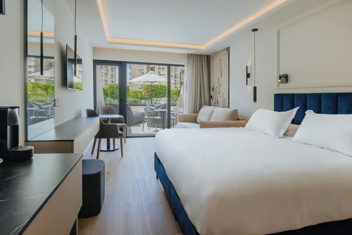Ein modernes Hotelzimmer mit einem großen Bett, einer Sitzecke mit Tisch und Stühlen sowie einem Balkon mit Gartenmöbeln.