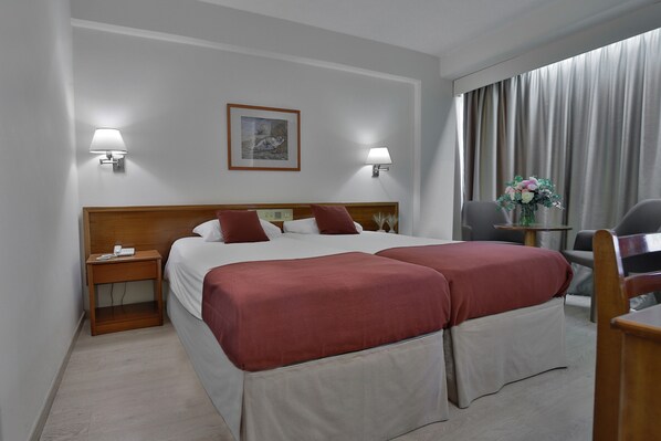 Habitación de hotel con cama doble, dos lámparas de noche, cabecero de madera, mesita de noche, escritorio con silla y un amplio ventanal con cortinas.