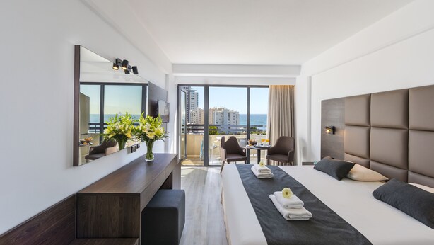 Habitación de hotel moderna con una cama grande, un escritorio, una silla y un balcón con vistas a la ciudad.