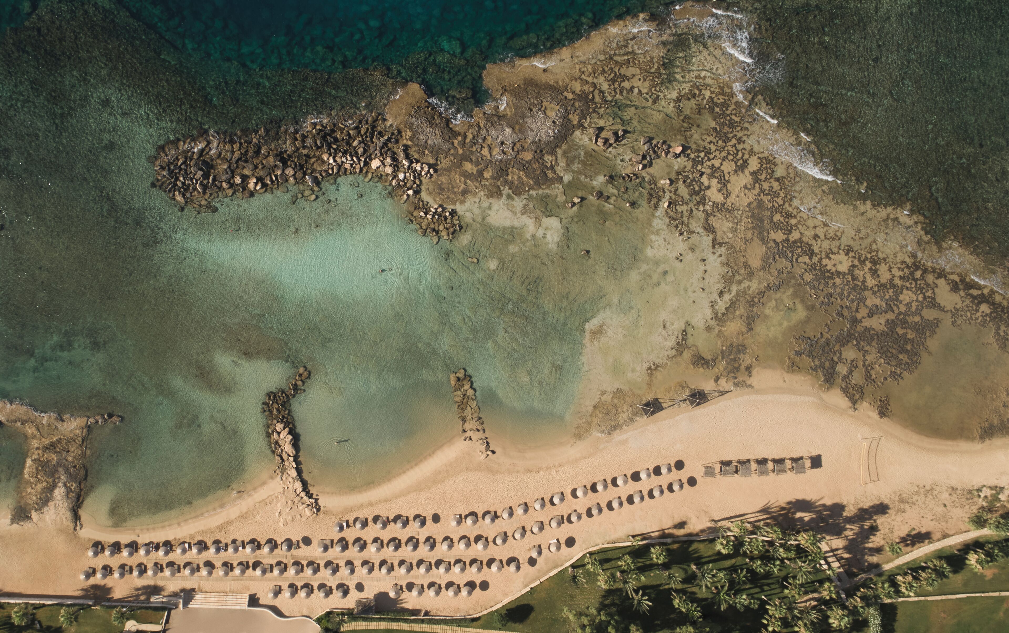Vista aerea di un resort sulla spiaggia con file di lettini circolari, una costa rocciosa e acque turchesi cristalline.