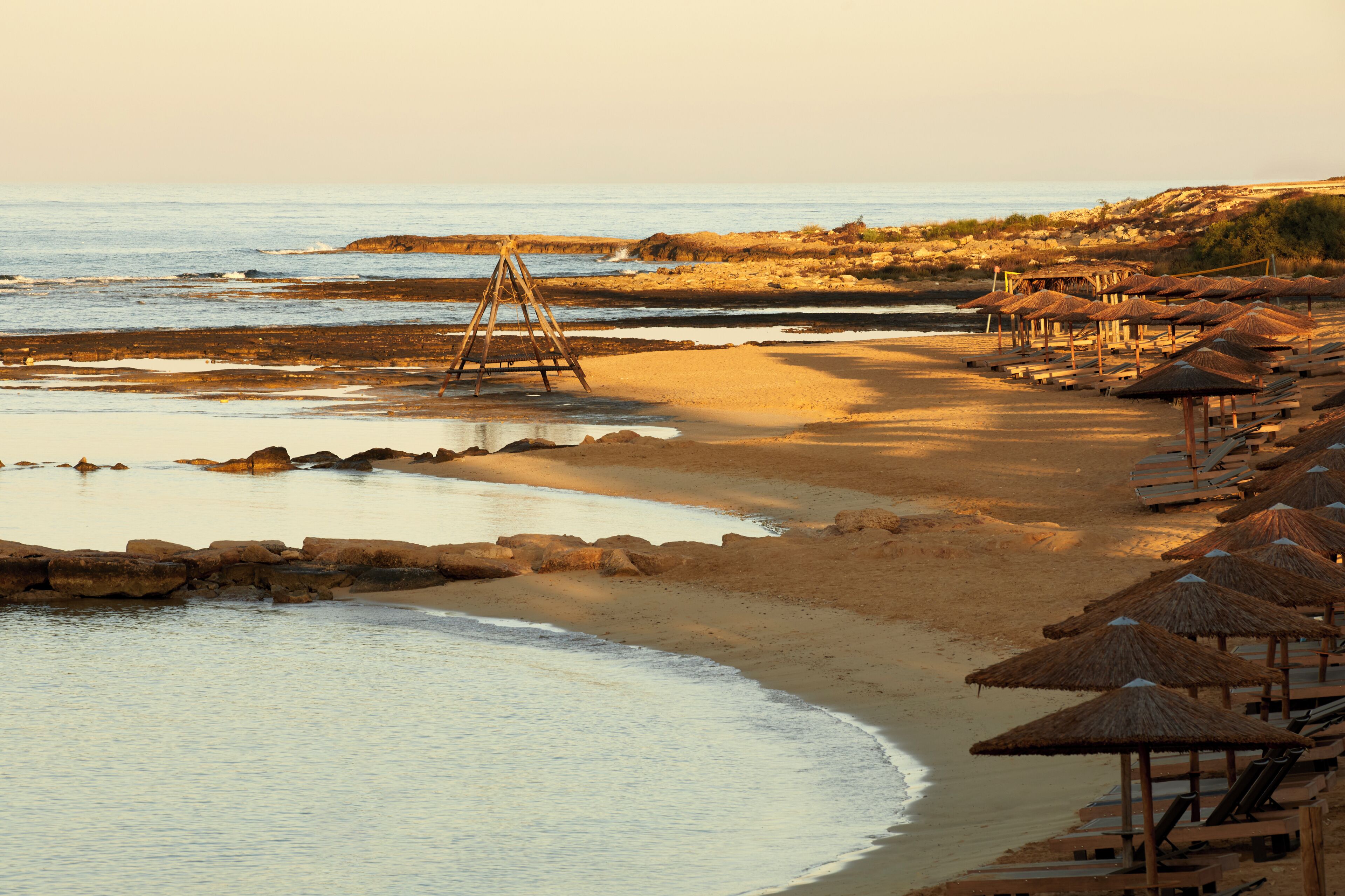 A beach with wooden loungers and thatched umbrellas, a swing set, and rocky outcrops.